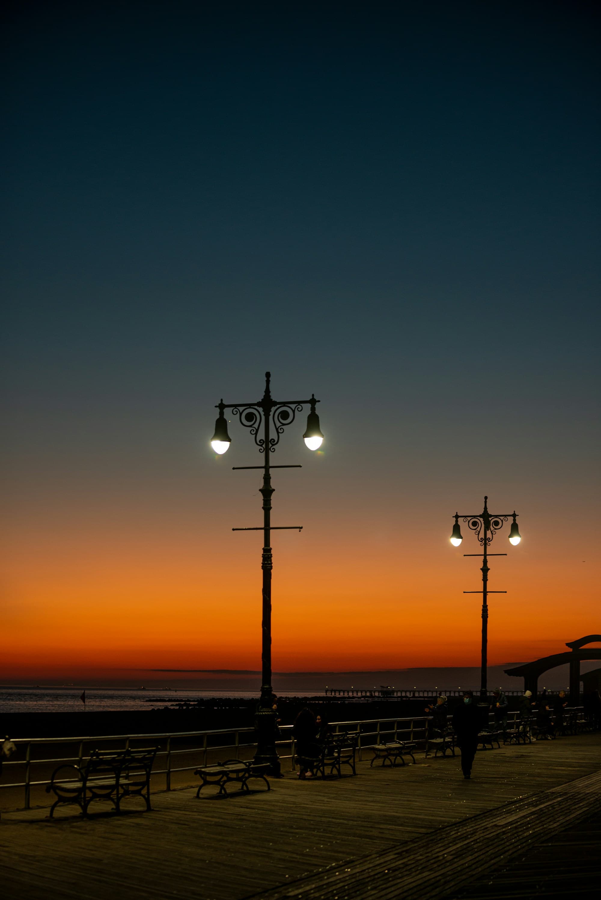 Glowing boardwalk lamps against an orange sunset over the ocean at Brighton Beach, Brooklyn.